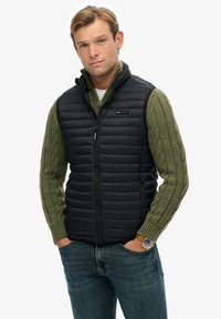 Chaleco acolchado negro con cuello alto, usado sobre un suéter verde de punto trenzado. Presenta bolsillos laterales y un logotipo de la marca en el pecho.