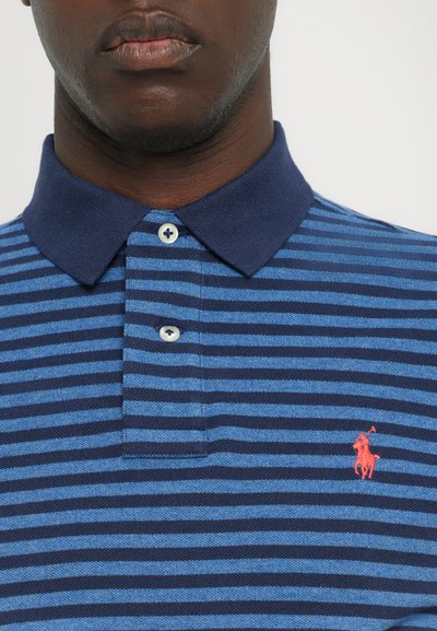 Jūras spēku un zilu strīpu polo krekls ar apkakles kakla izgriezumu, kuram ir mazs sarkans izšūts logo kreisajā krūtī un baltas pogas.