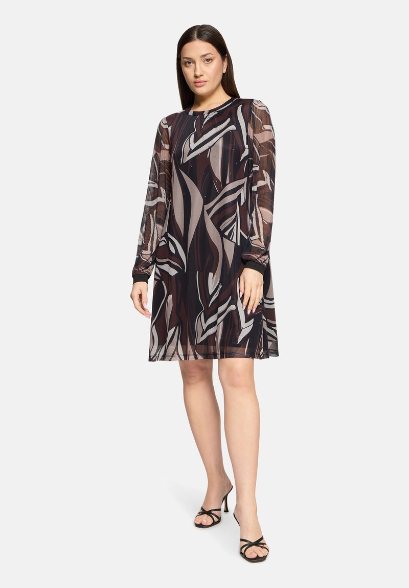 Woman wearing knee-length black dress with abstract beige and brown patterns and sheer long sleeves, paired with black strappy heels.