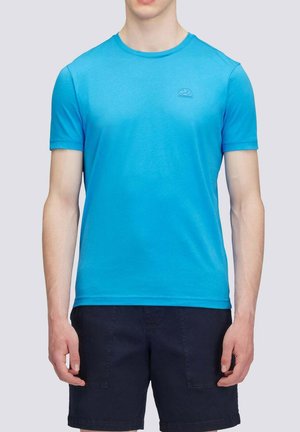 Uomo che indossa una t-shirt blu brillante a maniche corte e pantaloncini blu scuro, in piedi davanti a uno sfondo grigio chiaro uniforme.
