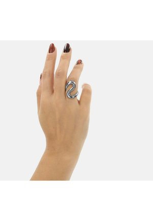 Dłoń z brązowymi i czarnymi paznokciami w wzory, nosząca srebrny pierścionek o abstrakcyjnym kształcie na środkowym palcu na białym tle.