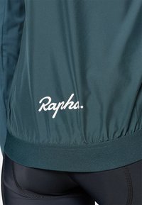 Turkusowa kurtka rowerowa z elastycznym dołem, z białym logo "Rapha." Gładka tekstura, lekki materiał i dopasowany krój.