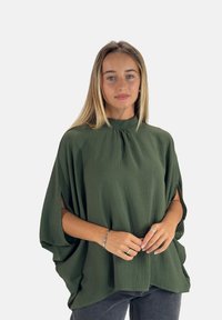 Blusa verde con tessuto testurizzato, collo alto, vestibilità ampia e maniche corte aperte. Abbinata a jeans scuri, che mostrano una silhouette rilassata.