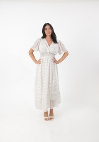 Abito maxi bianco a fantasia floreale con maniche corte a sbuffo e vita arricciata. Indossato con sandali con tacco leggero su uno sfondo neutro.