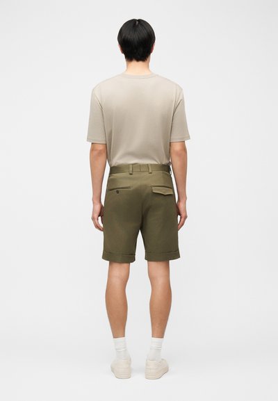 Άνδρας που φοράει μπεζ πουκάμισο με κοντά μανίκια μπουζωμένο σε ελιά πράσινη βερμούδα με πίσω τσέπες, λευκές κάλτσες και λευκά αθλητικά παπούτσια, γυρισμένος από την πλάτη σε απλό φόντο.