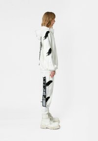 Chaqueta blanca con capucha y detalles de alas negras, combinada con pantalones a juego que presentan motivos de alas y acentos de etiqueta. Completado con botas blancas.