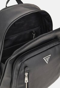 Zaino nero realizzato in materiale sintetico liscio, con un compartimento principale con zip, tasche interne e un dettaglio in metallo a forma triangolare.