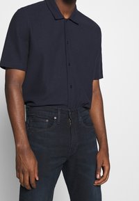 Marinblå pikétröja med korta ärmar, spread-krage, knappstängning och texturerat tyg, kombinerad med mörka denimjeans med dragkedja.