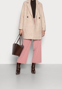 Béžový dvouřadý kabát se zapínáním na knoflíky a bočními kapsami, nošený přes přiléhavý tmavý top, doplněný o široké růžové kalhoty a texturovanou hnědou kabelku.