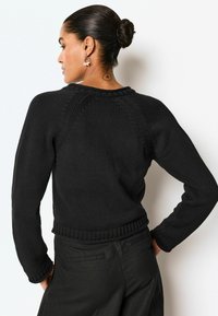 Sort strikket sweater med et struktureret mønster, rund halsudskæring og ribbet kant. Lange ærmer, der snævrer til ved håndleddene. Simpelt design, ingen synlige logoer.