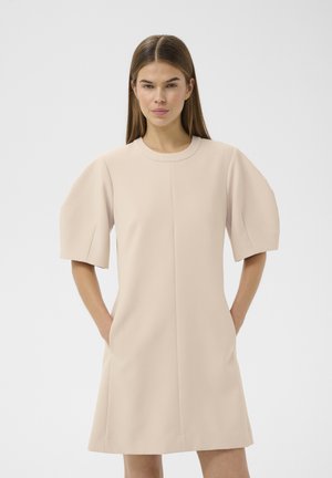 Vrouw draagt een beige jurk met korte, opgeblazen mouwen, staat met handen in de zakken tegen een effen lichtgrijze achtergrond.