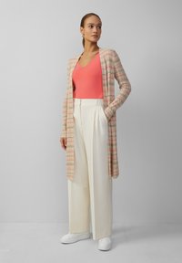 Langer, gemusterter Cardigan mit Wellenmuster in zartem Rosa und Creme über einem leuchtend korallenfarbenen Oberteil, kombiniert mit hoch taillierten weißen Hosen und weißen Sneakers.