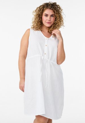 Kobieta o kręconych włosach, ubrana w białą sukienkę bez rękawów z guzikami i wiązanym w talii paskiem, pozująca z jedną ręką przy twarzy na białym tle.