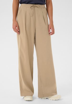 Beige wijde broek met taillekoord, gedragen met witte sneakers, getoond van taille tot vloer tegen een effen achtergrond.