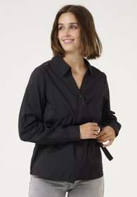 Camicia nera con bottoni, colletto, maniche lunghe e una vestibilità comoda. Il tessuto appare liscio con una leggera lucentezza e un dettaglio della tasca frontale.