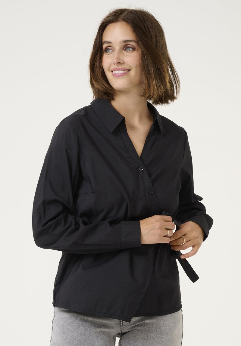 Camicia nera con bottoni, colletto, maniche lunghe e una vestibilità comoda. Il tessuto appare liscio con una leggera lucentezza e un dettaglio della tasca frontale.
