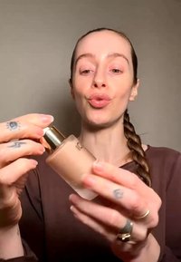 Young woman with braided hair holding and opening a frosted glass bottle with gold cap, likely a cosmetic product, against a plain background.