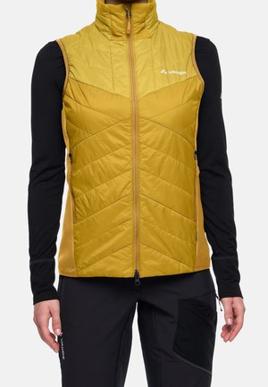 Persoon draagt een mosterdgele doorgestikte mouwloze vest over een zwart shirt met lange mouwen en een zwarte broek met een zak op het dijbeen.