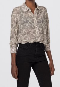 Camicia leggera button-up di colore beige con motivo fogliare nero. Presenta un colletto, maniche lunghe e tessuto testurizzato. Abbinata a jeans neri.