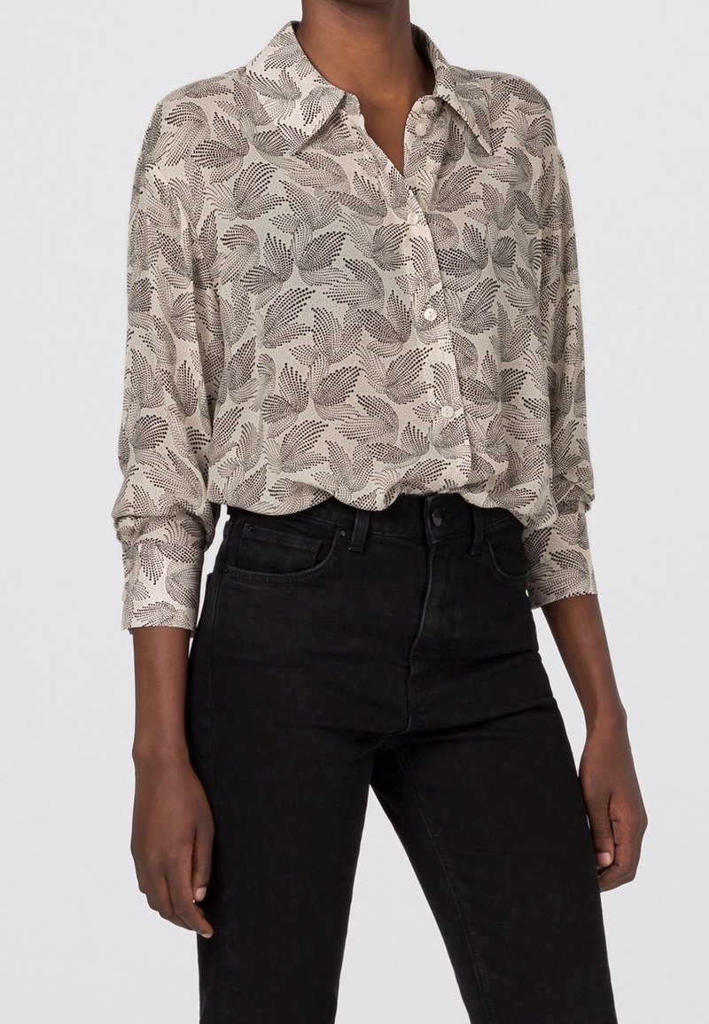 Camicia leggera button-up di colore beige con motivo fogliare nero. Presenta un colletto, maniche lunghe e tessuto testurizzato. Abbinata a jeans neri.