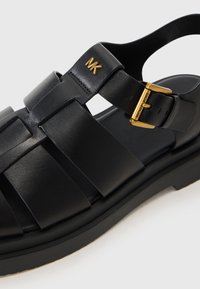 Sorte lær sandaler med kryssdesign, justerbar gullfarget spenne og en teksturert svart gummisåle. Gullfarget logo-akkent synlig.