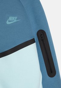 Sweatshirt da Nike com um tecido azul texturizado, um painel azul claro, uma faixa de destaque preta e um bolso com zíper na manga.