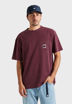 Homme portant un t-shirt oversize rouge foncé avec un logo sur la poche, un jean bleu clair avec une lanière noire et une casquette bleu marine avec un logo assorti.