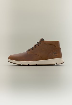 Brauner Lederknöchelstiefel mit schwarzen Schnürsenkeln, weißer Sohle und einem Baum-Logo an der Ferse, gezeigt auf einem schlichten hellen Hintergrund.