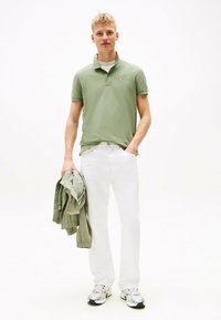 Polo de color verde claro, jeans blancos, sosteniendo una chaqueta verde a juego. Zapatillas con acentos metálicos. Atuendo simple y casual.