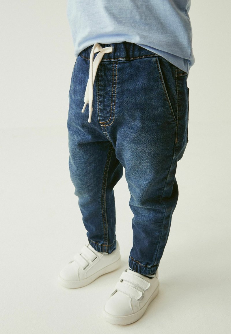 Enfant portant un pantalon jogger en denim bleu avec chevilles élastiques et des baskets blanches à bandes velcro sur fond clair.