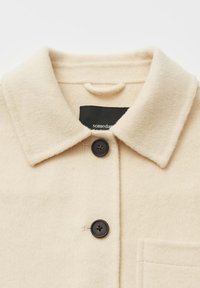 Giacca beige in misto lana con colletto a punta, due bottoni neri e una tasca frontale; tessuto liscio, finitura morbida.