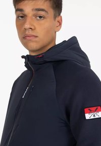 Chaqueta con capucha azul marino en un tejido suave, con un parche rojo y blanco en la manga izquierda y detalles con cordones en el cuello.