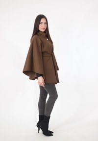 Bruine cape-jas met ceintuur, hoge kraag en losse mouwen, gecombineerd met grijze skinny jeans en zwarte enkellaarzen.