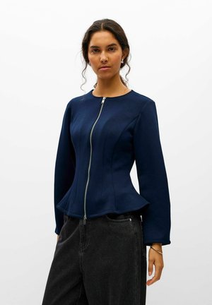 Modèle portant une veste ajustée bleu marine, avec une fermeture éclair sur le devant et un bas évasé, associée à un jean noir sur un fond blanc uni.