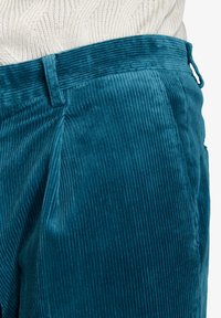 Teal corduroy-housut, joissa on pystysuora ribbitekstuuri, litteä vyötärö ja sivusaumat. Kangas näyttää pehmeältä ja ylelliseltä.