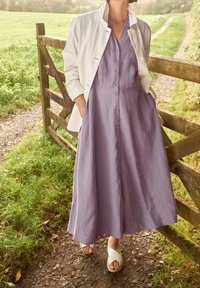 Lawendowa sukienka z lnu z guzikami z przodu, w kształcie litery A, z bocznymi kieszeniami, w zestawieniu z białą kurtką i jasnymi sandałami.