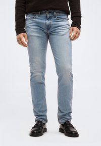 Άνδρας που φοράει ανοιχτό μπλε τζιν στενής γραμμής, μαύρα παπούτσια τύπου loafer και σκούρο καφέ πουκάμισο με μακριά μανίκια, στέκεται μπροστά σε απλό φόντο.