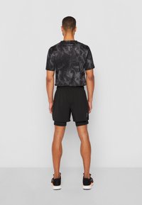 Mies, joka seisoo selkä kameraan päin, päällä musta savumallinen urheilupaita, kerrosmustat shortsit ja mustat juoksukengät oranssilla pohjalla.