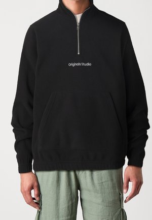 Personne portant un sweatshirt noir à demi-zippé avec le texte "originals Studio" et un pantalon cargo vert clair, se tenant devant un fond uni.