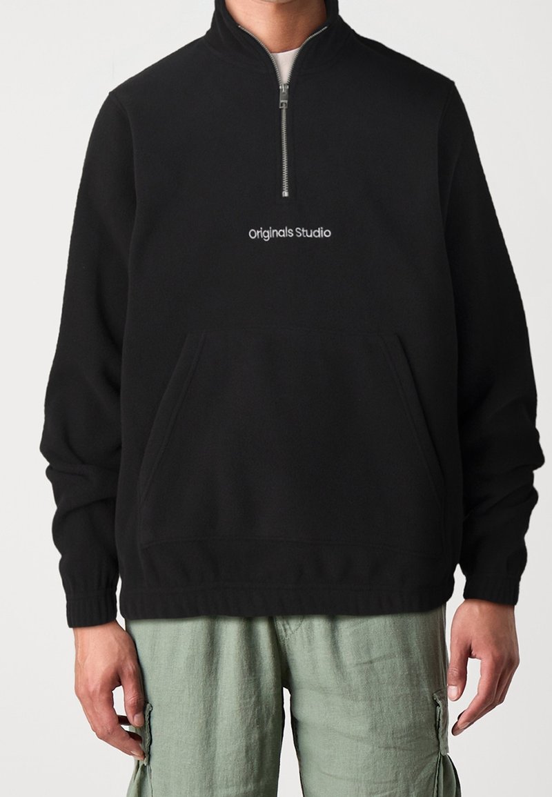 Personne portant un sweatshirt noir à demi-zippé avec le texte "originals Studio" et un pantalon cargo vert clair, se tenant devant un fond uni.