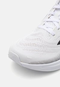 Gros plan sur la pointe d'une chaussure de sport blanche en matériel mesh respirant avec une semelle texturée sur un fond uni.