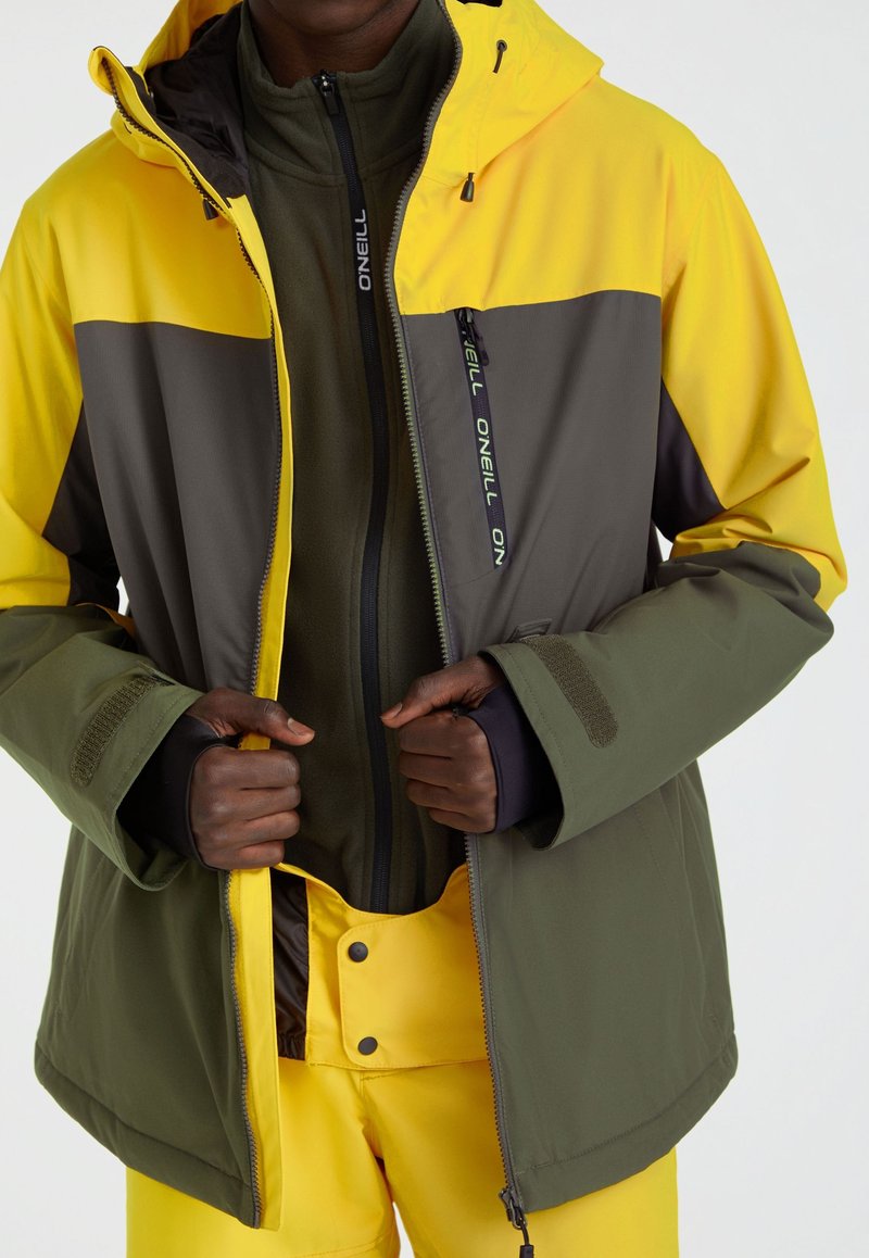 Kurtka w bloki kolorystyczne z panelami w kolorach żółtym, szarym i oliwkowym. Miękka tkanina, zamek z przodu i mankiety na rzepy. Nosi się ją na polarowej bluzie zapinanej na zamek.