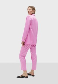 Pink satin suit with a loose-fitting blazer and tapered pants. Black slingback shoes with a textured upper. Simple gold button accents.