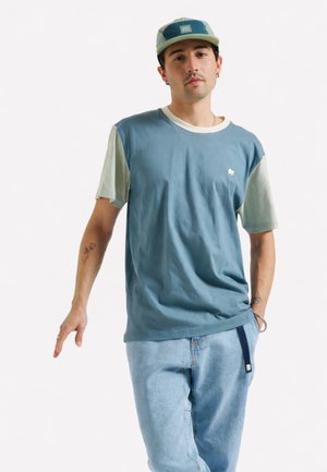 Jeune homme portant une chemise à manches courtes bleue et beige, un jean bleu clair et une casquette patchwork bleue et verte, posant devant un fond blanc.