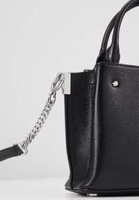 Sac à main en cuir noir texturé avec une bandoulière chaîne argentée attachée à un fermoir métallique sur fond neutre.