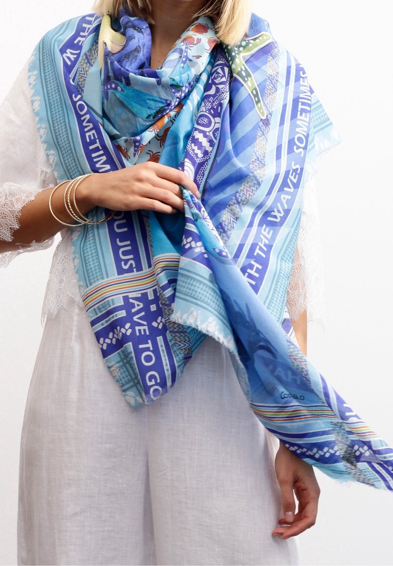 Femme portant une écharpe bleue colorée à motifs sur une tenue blanche bordée de dentelle, tenant un côté de l'écharpe avec des bracelets au poignet.