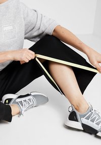 Zwarte zippered joggers met een gele rits, gecombineerd met grijze en zwarte hardloopschoenen met een geavanceerde mesh bovenkant en een witte zool.