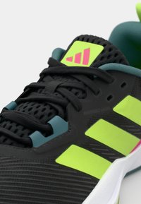 Black athletic shoe with mesh upper, green accents, and pink details. Features include adjustable lacing and three prominent side stripes.