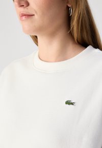 White sweatshirt with a textured finish, featuring a small green embroidered crocodile logo on the left chest and a classic round neckline.