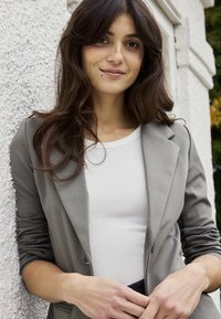 Młoda kobieta o długich ciemnych włosach, ubrana w szarą marynarkę na białym topie, stoi na zewnątrz przy białej teksturowanej ścianie.